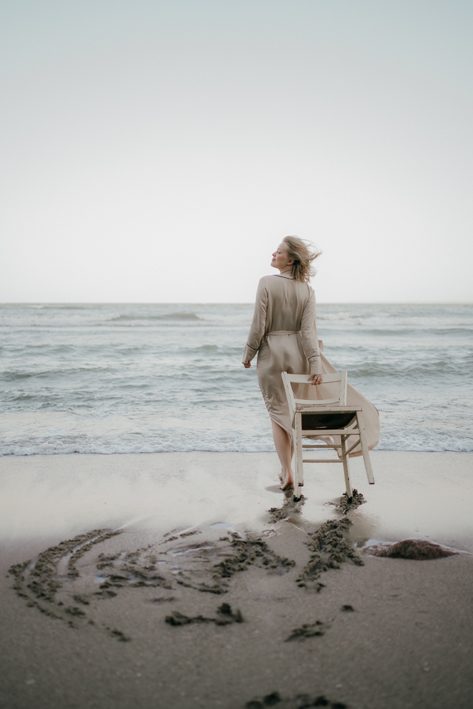 Frau in beigem Kleid steht mit Stuhl am Strand Grömitz. 
Elegante Frau am Meer mit wehendem Haar.